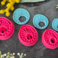 Three 3d printed polymer clay cutters in bright pink, arranged on a dark textured surface surrounded by colorful flowers. Halo Beam Polymer Clay Cutter