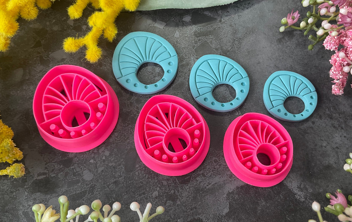 Three 3d printed polymer clay cutters in bright pink, arranged on a dark textured surface surrounded by colorful flowers. Halo Beam Polymer Clay Cutter
