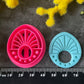 Three 3d printed polymer clay cutters in bright pink, arranged on a dark textured surface surrounded by colorful flowers. Halo Beam Polymer Clay Cutter