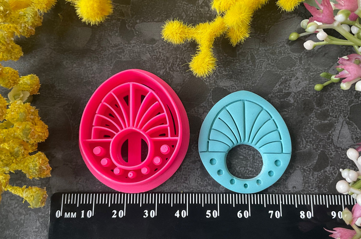 Three 3d printed polymer clay cutters in bright pink, arranged on a dark textured surface surrounded by colorful flowers. Halo Beam Polymer Clay Cutter