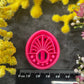 Three 3d printed polymer clay cutters in bright pink, arranged on a dark textured surface surrounded by colorful flowers. Halo Beam Polymer Clay Cutter