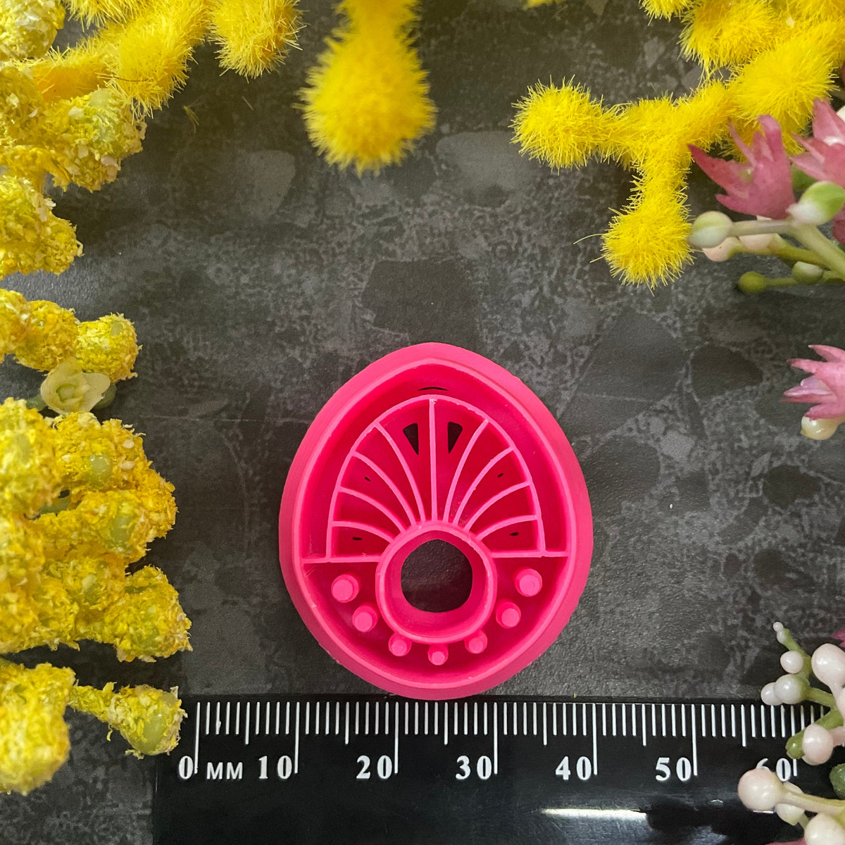 Three 3d printed polymer clay cutters in bright pink, arranged on a dark textured surface surrounded by colorful flowers. Halo Beam Polymer Clay Cutter