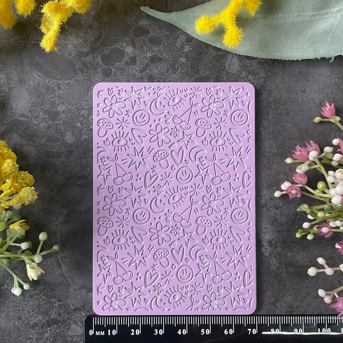 A lavender purple silicone baking mold with embossed floral and heart patterns, displayed on a dark gray textured surface with a black ruler at the bottom for scale, surrounded by yellow and pink flowers.