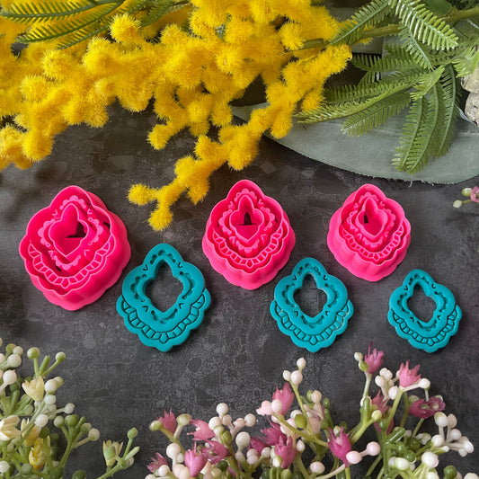 Three hot pink 3d printed polymer clay  cutters arranged on a dark textured surface with decorative flowers around them, set against yellow mimosa and pink/white blossoms. Heirloom Lace Polymer Clay Cutter