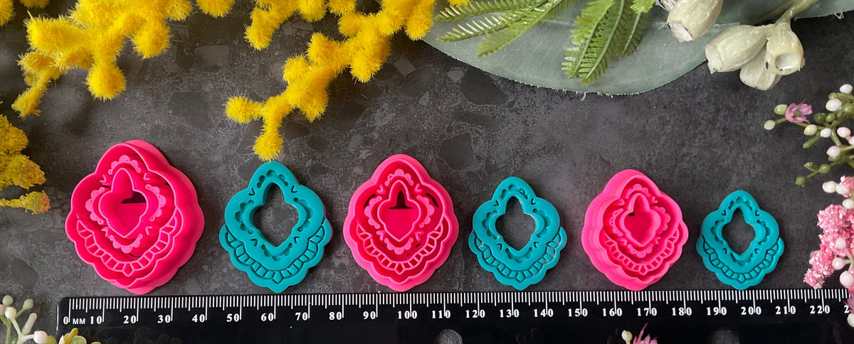 Three hot pink 3d printed polymer clay  cutters arranged on a dark textured surface with decorative flowers around them, set against yellow mimosa and pink/white blossoms. Heirloom Lace Polymer Clay Cutter