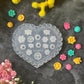 A heart-shaped clear plastic mold with floral and geometric engravings, surrounded by small colorful flower-shaped beads or charms on a dark textured surface with yellow and pink flowers.