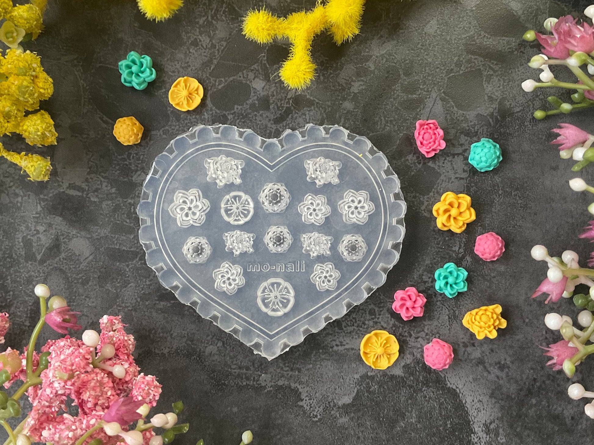 A heart-shaped clear plastic mold with floral and geometric engravings, surrounded by small colorful flower-shaped beads or charms on a dark textured surface with yellow and pink flowers.