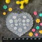 A heart-shaped clear plastic mold with white floral patterns, displayed on a dark textured surface surrounded by yellow, pink, and green flowers, with a black ruler at the bottom showing measurements.