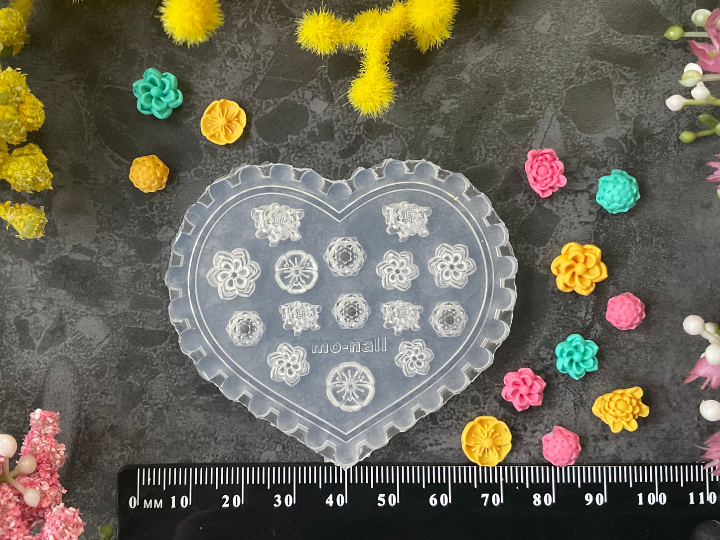 A heart-shaped clear plastic mold with white floral patterns, displayed on a dark textured surface surrounded by yellow, pink, and green flowers, with a black ruler at the bottom showing measurements.