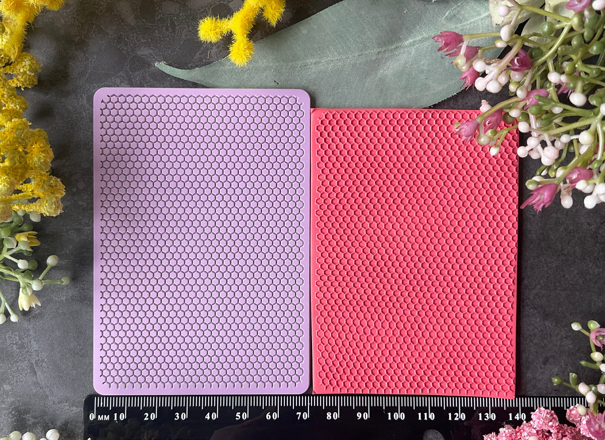 Two textured rectangular mats, one light purple and one coral pink, displayed side-by-side on a dark gray surface with a black measuring ruler at the bottom and yellow and pink flowers framing the scene.