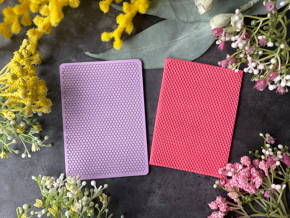 Two textured rectangular cards or cases, one lavender purple and one coral pink, displayed on a dark gray surface surrounded by yellow and pink flowers.