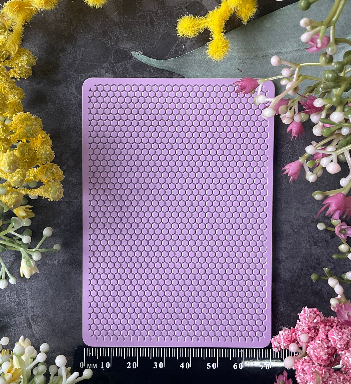 A rectangular lavender purple silicone mat with a raised honeycomb pattern, shown flat-lay on a dark gray surface surrounded by yellow and pink flowers, with a black ruler at the bottom indicating scale.