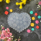 A heart-shaped clear plastic craft mold with embossed floral patterns, displayed on a dark textured surface surrounded by colorful flower petals and artificial yellow and pink flowers.