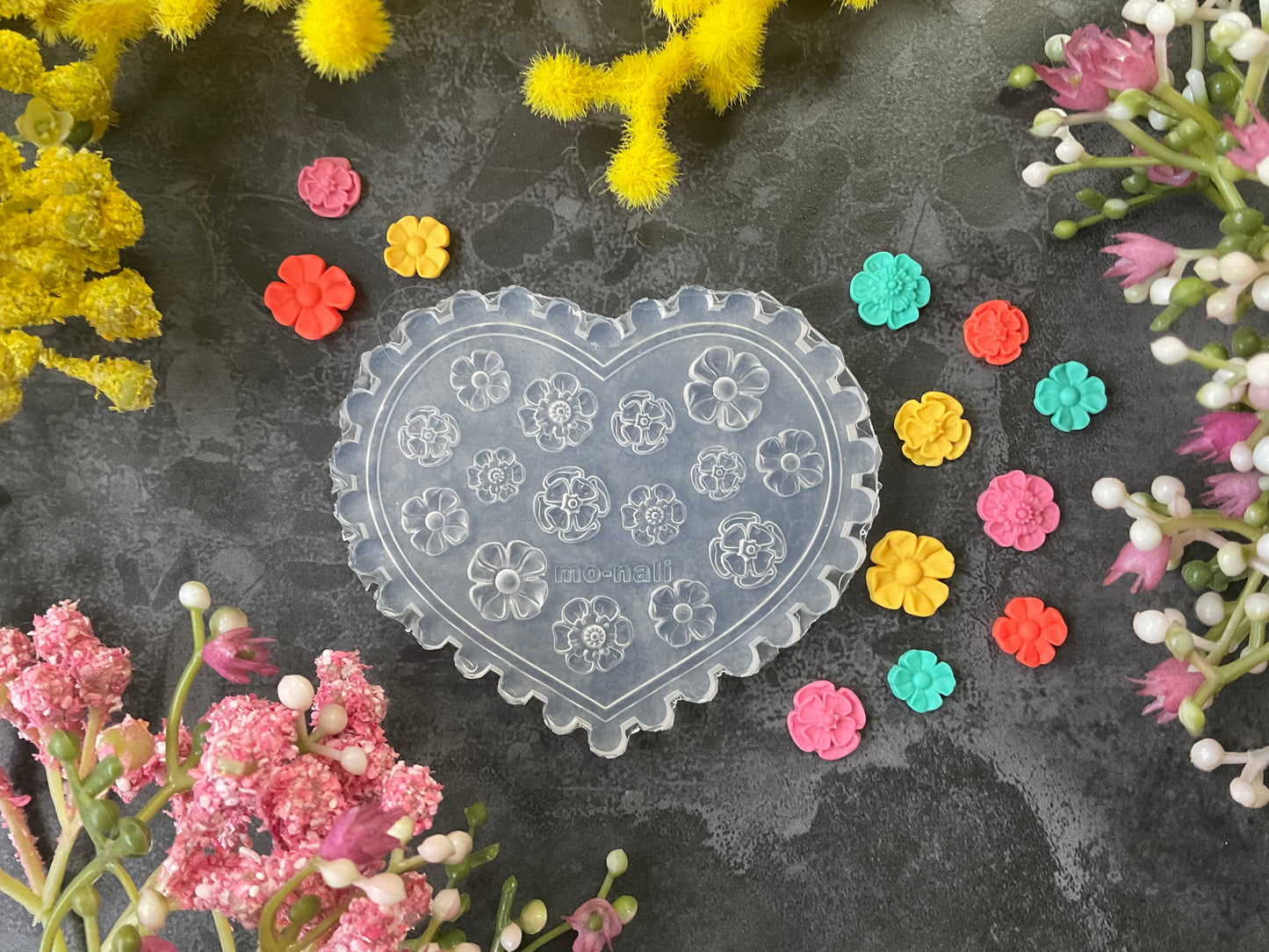 A heart-shaped clear plastic craft mold with embossed floral patterns, displayed on a dark textured surface surrounded by colorful flower petals and artificial yellow and pink flowers.