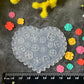 A heart-shaped clear plastic craft mold with floral recessed patterns, displayed on a dark textured surface with a black ruler at the bottom for scale, surrounded by small colorful flower cutouts and faux flowers.