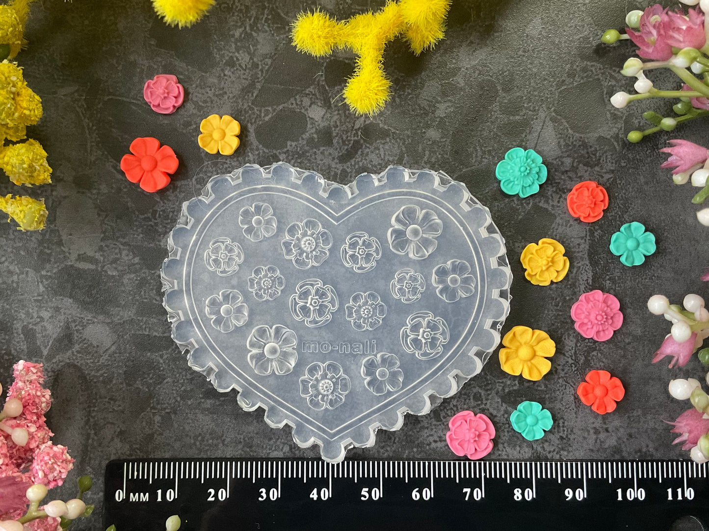 A heart-shaped clear plastic craft mold with floral recessed patterns, displayed on a dark textured surface with a black ruler at the bottom for scale, surrounded by small colorful flower cutouts and faux flowers.