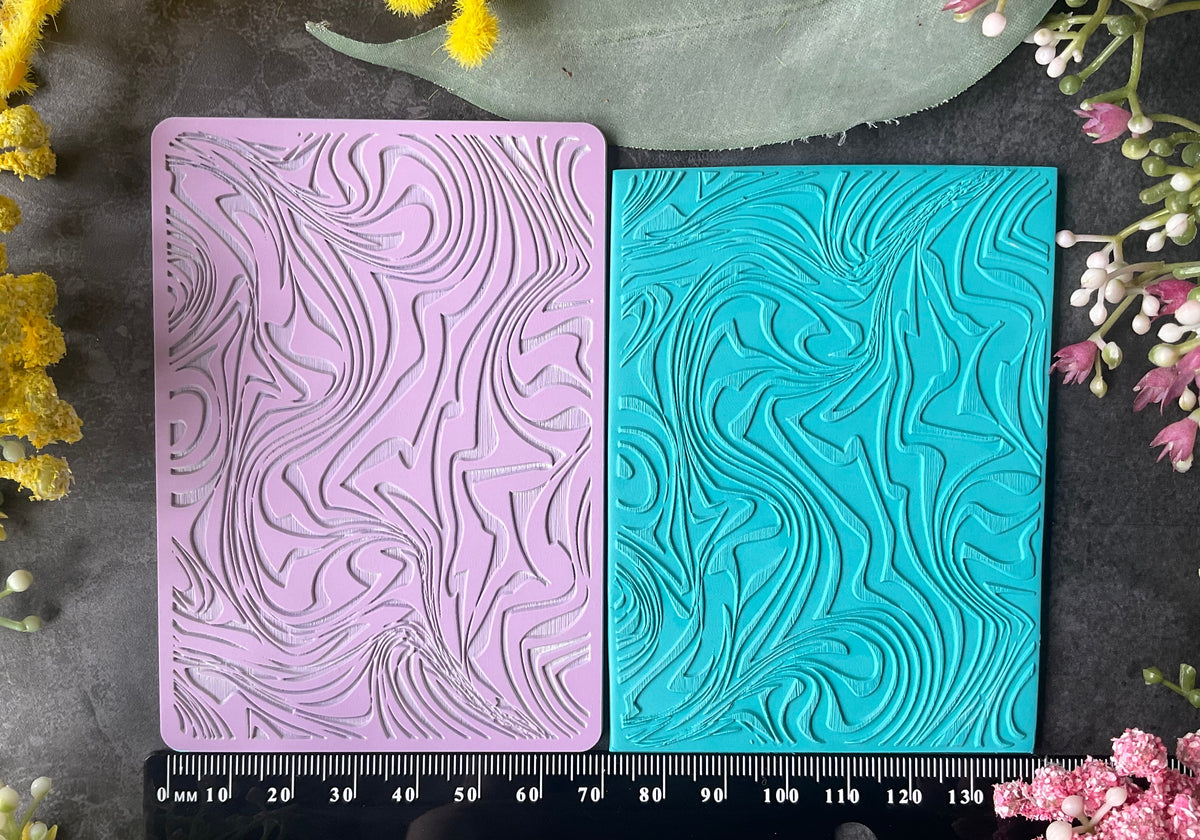 Two rectangular embossed rubber stamps in lavender pink and turquoise blue Ice-cream Swirl Texture Mat texture, displayed side-by-side on a dark textured surface with a black ruler at the bottom and floral decorations around the edges.