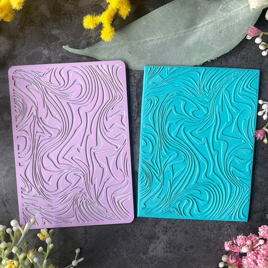 Two rectangular embossed stamp blocks or cards in lavender purple and turquoise blue Ice-cream Swirl Texture Mat, placed side by side on a dark textured surface with decorative flowers around the edges.