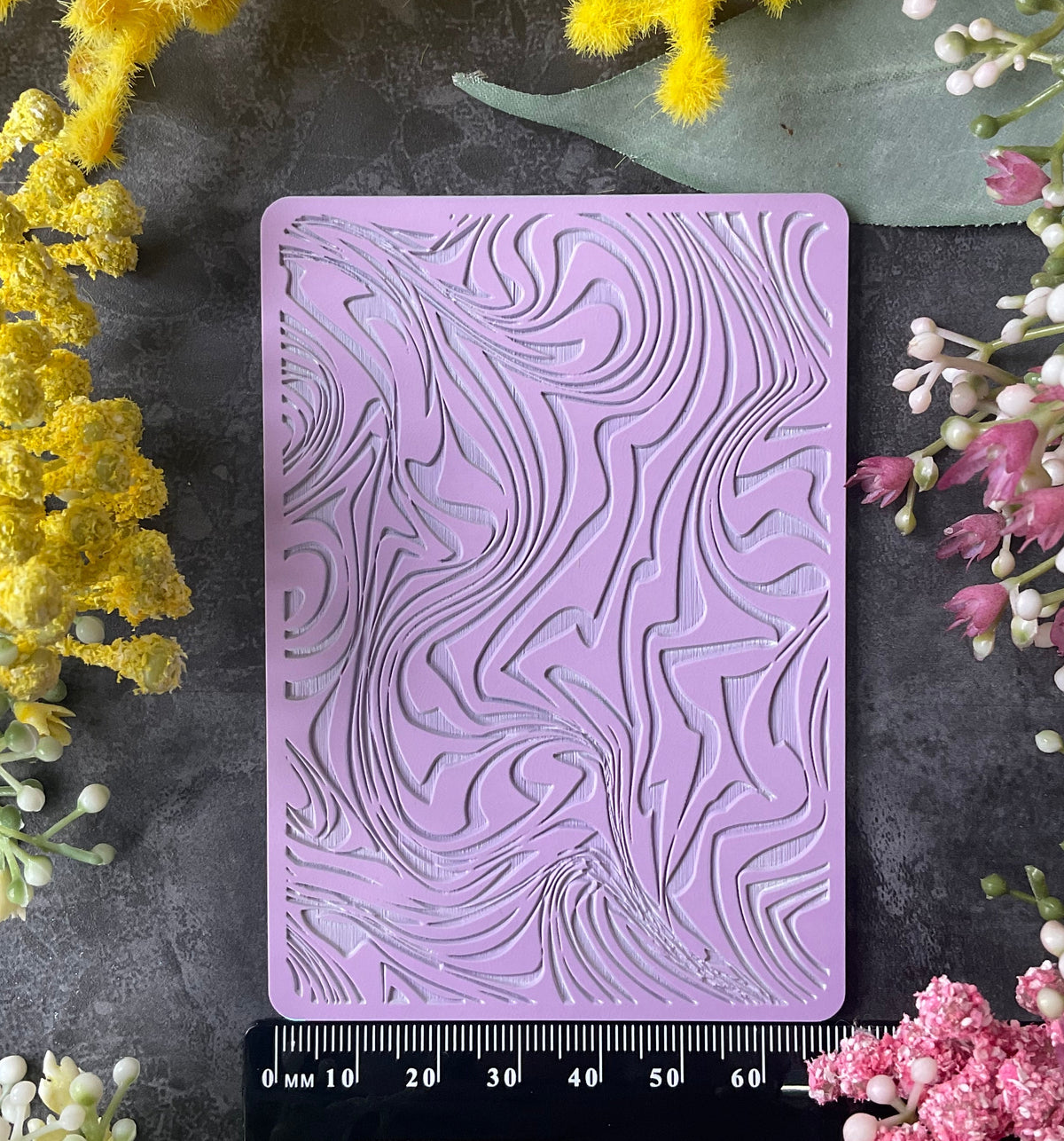 A lavender purple silicone mold with an embossed Ice-cream Swirl Texture Mat wavy wood grain pattern, displayed on a dark gray textured surface with a black ruler at the bottom and yellow and pink flowers framing the product.