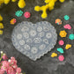 A heart-shaped clear plastic mold with floral and geometric patterns, surrounded by small colorful flower-shaped embellishments and surrounded by yellow and pink flowers on a dark textured background.
