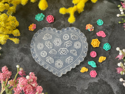 A heart-shaped clear plastic mold with floral and geometric patterns, surrounded by small colorful flower-shaped embellishments and surrounded by yellow and pink flowers on a dark textured background.
