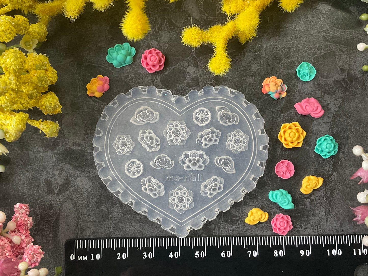 A heart-shaped clear plastic craft mold with floral and crescent moon cutouts, displayed on a dark textured surface surrounded by yellow and pink flowers, small colorful flower pieces, and a black ruler at the bottom.