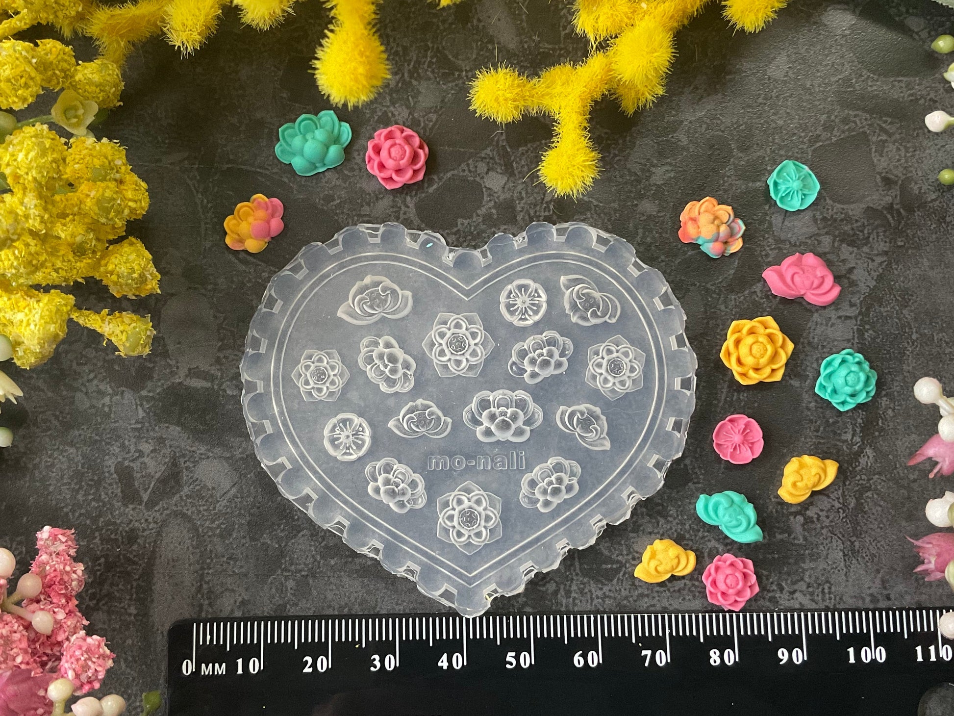 A heart-shaped clear plastic craft mold with floral and crescent moon cutouts, displayed on a dark textured surface surrounded by yellow and pink flowers, small colorful flower pieces, and a black ruler at the bottom.