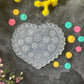 A heart-shaped clear plastic mold with floral patterns, displayed on a dark textured surface surrounded by yellow, pink, and teal flowers.