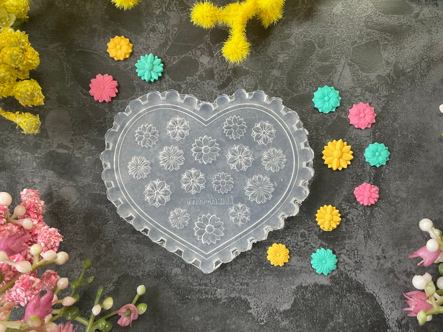 A heart-shaped clear plastic mold with floral patterns, displayed on a dark textured surface surrounded by yellow, pink, and teal flowers.