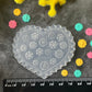 A heart-shaped clear plastic mold with floral snowflake-like patterns, displayed on a dark textured surface with a black ruler at the bottom for scale, surrounded by small colorful flower-shaped embellishments and yellow/pink floral decorations.