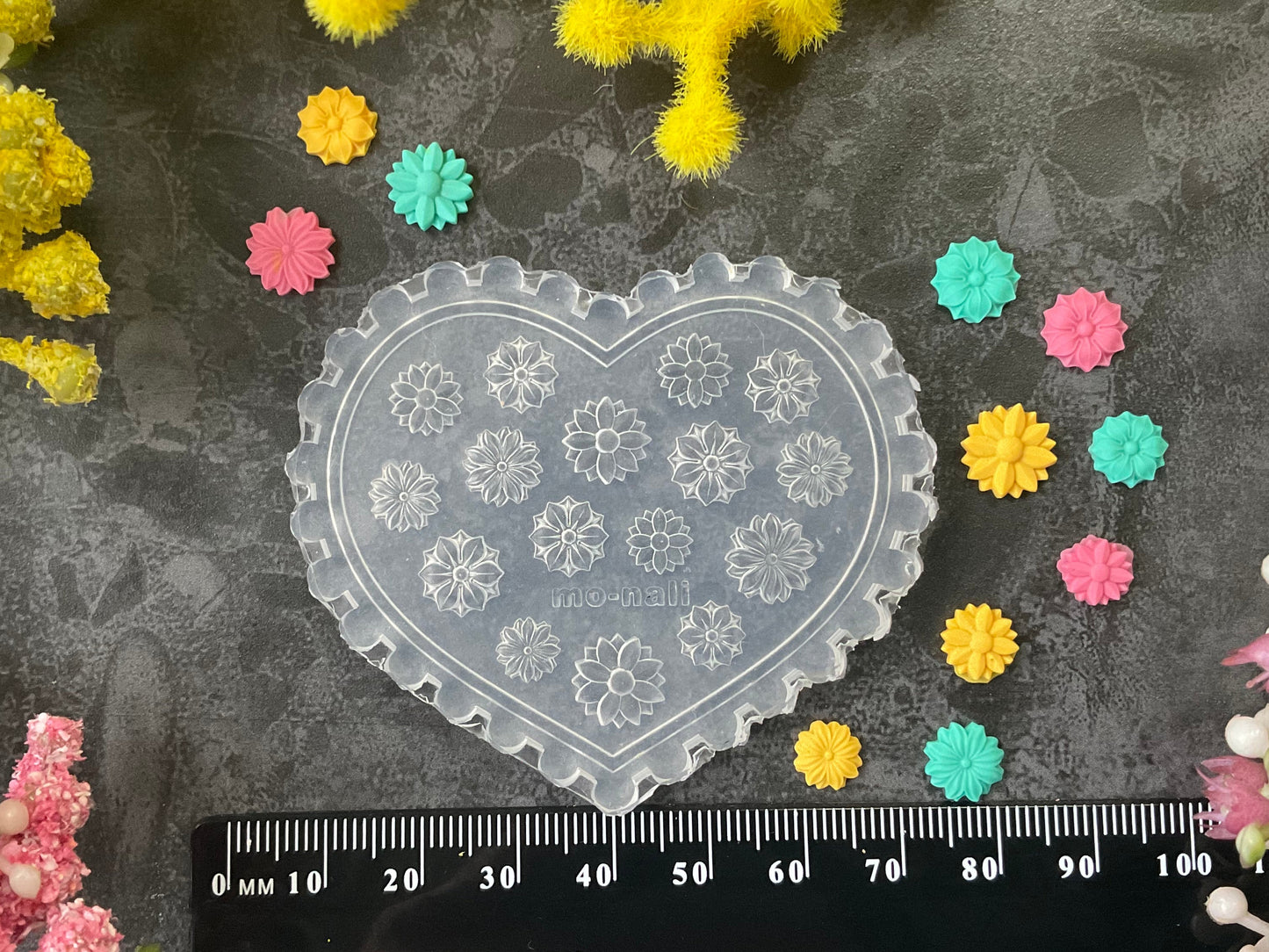 A heart-shaped clear plastic mold with floral snowflake-like patterns, displayed on a dark textured surface with a black ruler at the bottom for scale, surrounded by small colorful flower-shaped embellishments and yellow/pink floral decorations.