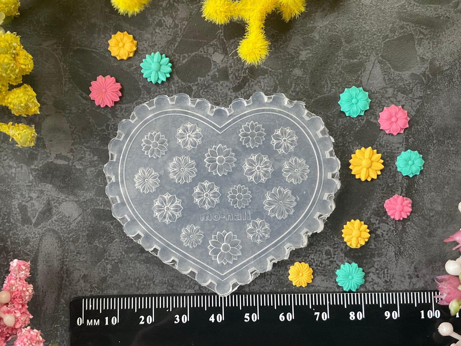 A heart-shaped clear plastic mold with floral snowflake-like patterns, displayed on a dark textured surface with a black ruler at the bottom for scale, surrounded by small colorful flower-shaped embellishments and yellow/pink floral decorations.