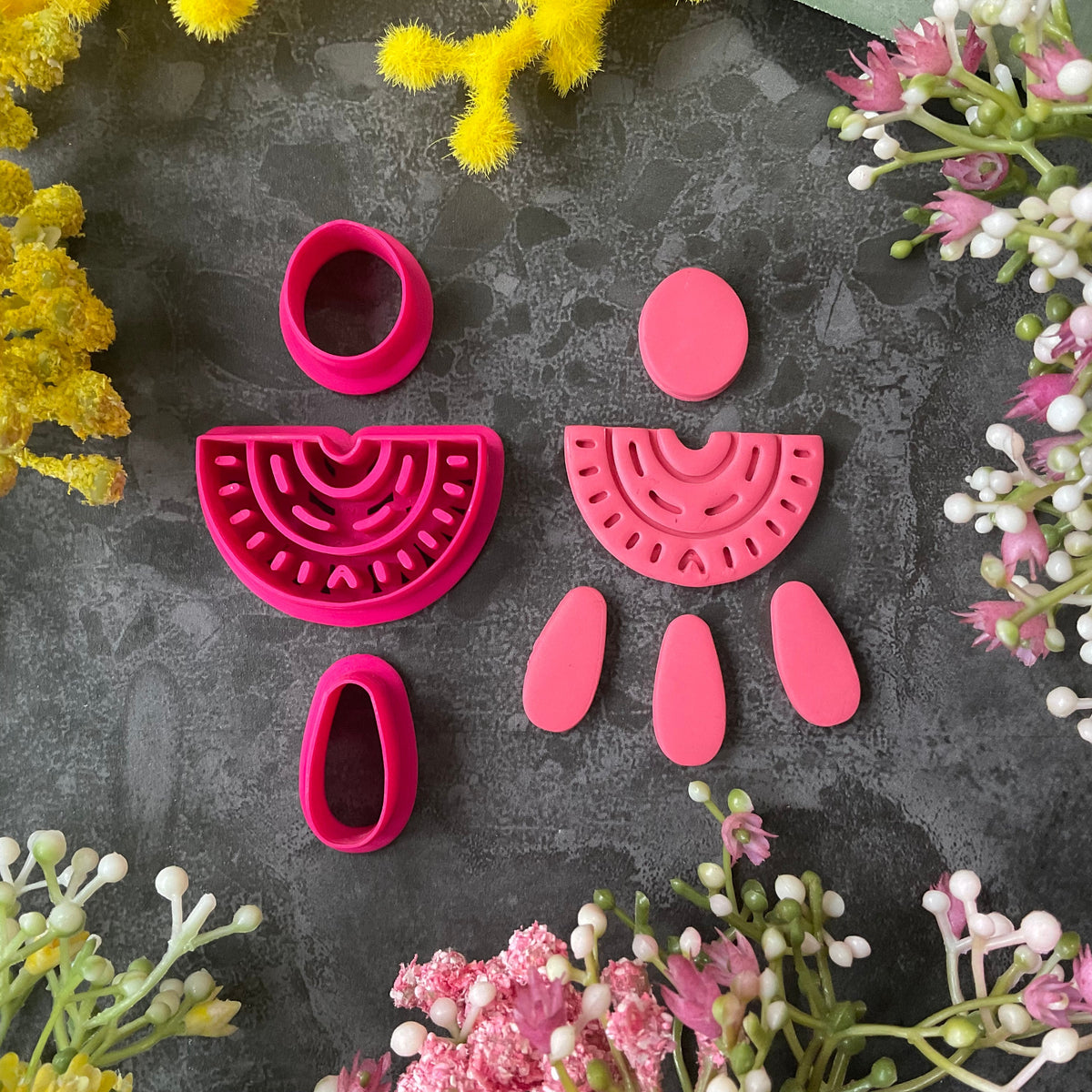 A set of bright pink 3d printed polymer clay cutters arranged on a dark textured surface, surrounded by yellow and pink flowers. The set includes two semicircle cutters with different line patterns, two oval cutters, and two teardrop-shaped cutters. Nomad Arch Polymer Clay Cutter Set.