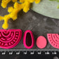A set of bright pink 3d printed polymer clay cutters arranged on a dark textured surface, surrounded by yellow and pink flowers. The set includes two semicircle cutters with different line patterns, two oval cutters, and two teardrop-shaped cutters. Nomad Arch Polymer Clay Cutter Set.