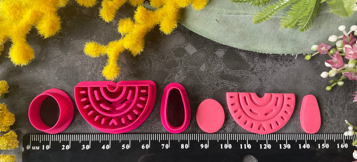 A set of bright pink 3d printed polymer clay cutters arranged on a dark textured surface, surrounded by yellow and pink flowers. The set includes two semicircle cutters with different line patterns, two oval cutters, and two teardrop-shaped cutters. Nomad Arch Polymer Clay Cutter Set.