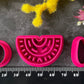 A set of bright pink 3d printed polymer clay cutters arranged on a dark textured surface, surrounded by yellow and pink flowers. The set includes two semicircle cutters with different line patterns, two oval cutters, and two teardrop-shaped cutters. Nomad Arch Polymer Clay Cutter Set.