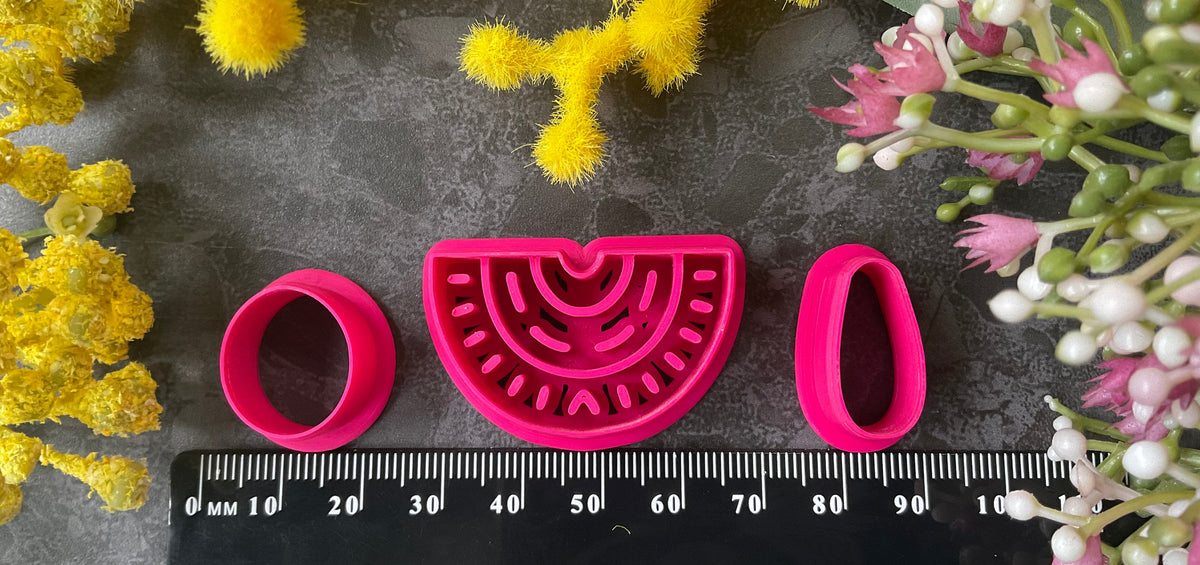 A set of bright pink 3d printed polymer clay cutters arranged on a dark textured surface, surrounded by yellow and pink flowers. The set includes two semicircle cutters with different line patterns, two oval cutters, and two teardrop-shaped cutters. Nomad Arch Polymer Clay Cutter Set.