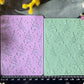 Two rectangular craft stamp blocks, one lavender purple and one mint green, both embossed with a repeating Christmas tree pattern, displayed on a dark textured surface with a black ruler at the bottom for scale and surrounded by yellow and pink flowers.