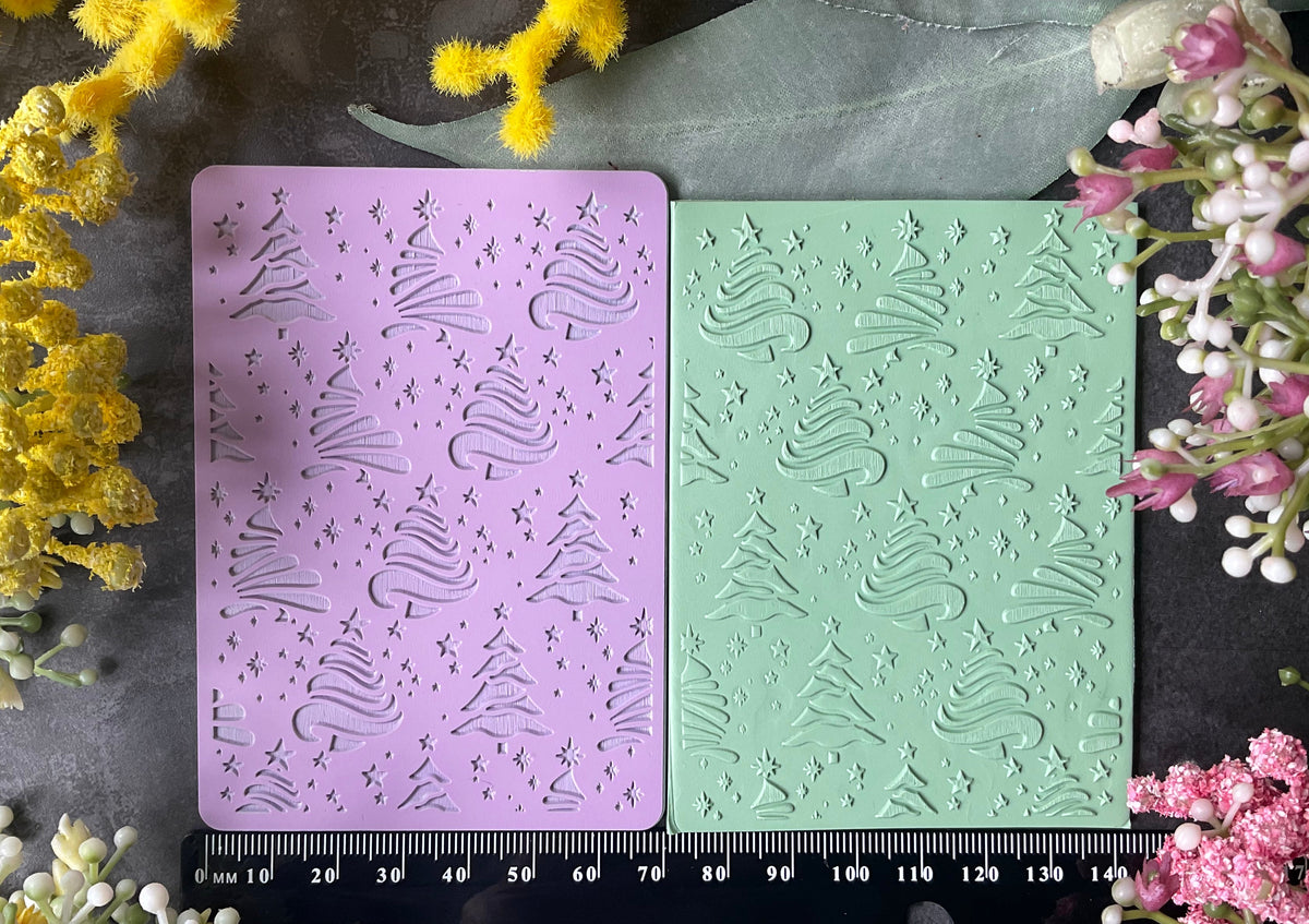 Two rectangular craft stamp blocks, one lavender purple and one mint green, both embossed with a repeating Christmas tree pattern, displayed on a dark textured surface with a black ruler at the bottom for scale and surrounded by yellow and pink flowers.