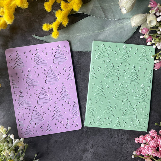 Two rectangular embossed cards or stamps in pastel purple and mint green, decorated with a Christmas tree and swirl patterns, arranged on a dark textured surface with yellow and pink flowers around them.