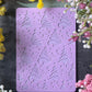 A lavender purple rectangular metal stamp with embossed Christmas tree and snowflake patterns, displayed on a dark textured surface and surrounded by yellow and pink flowers with a black ruler at the bottom for scale.