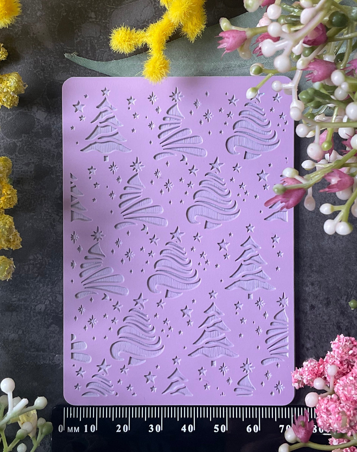 A lavender purple rectangular metal stamp with embossed Christmas tree and snowflake patterns, displayed on a dark textured surface and surrounded by yellow and pink flowers with a black ruler at the bottom for scale.