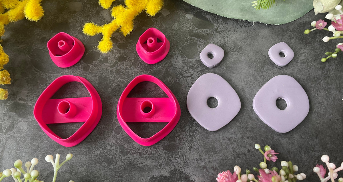 A flat-lay of polymer clay pieces and 3d printed polymer clay cutters on a dark gray textured surface, surrounded by decorative yellow, green, and pink flowers. The pieces include two bright magenta-pink geometric shaped polymer clay cutters and two light lavender-gray organic shapes, with smaller matching spacers. Organic Pebble Petal Polymer Clay Cutter Set