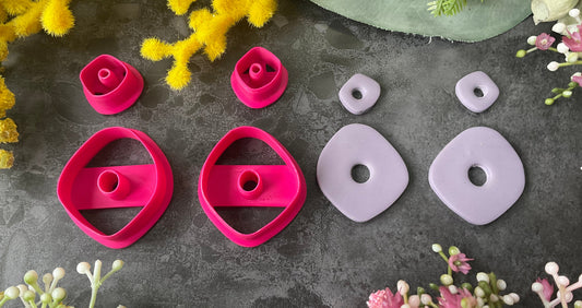 A flat-lay of polymer clay pieces and 3d printed polymer clay cutters on a dark gray textured surface, surrounded by decorative yellow, green, and pink flowers. The pieces include two bright magenta-pink geometric shaped polymer clay cutters and two light lavender-gray organic shapes, with smaller matching spacers. Organic Pebble Petal Polymer Clay Cutter Set