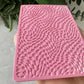 Pink textured square object held by a hand with a blurred green plant background