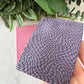 A close-up of a textured rectangular sample showing a woven or basketweave pattern in a muted lavender-purple color, with a pink sample partially visible behind it. A hand is holding the front sample, and green leaves are placed around the top edge against a light neutral background.