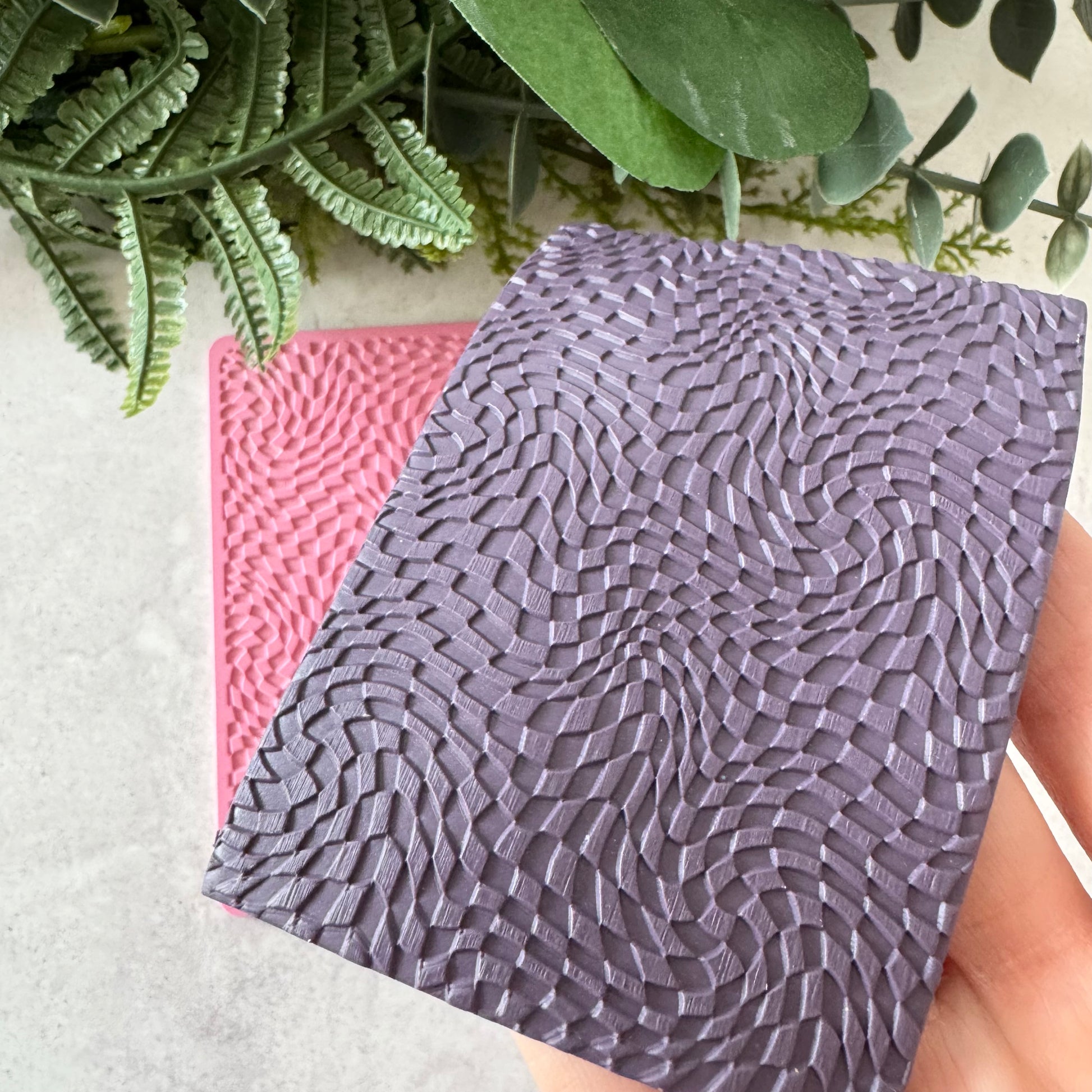 A close-up of a textured rectangular sample showing a woven or basketweave pattern in a muted lavender-purple color, with a pink sample partially visible behind it. A hand is holding the front sample, and green leaves are placed around the top edge against a light neutral background.