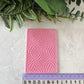 Pink embossed rectangular object on a light gray surface with green plants in the background