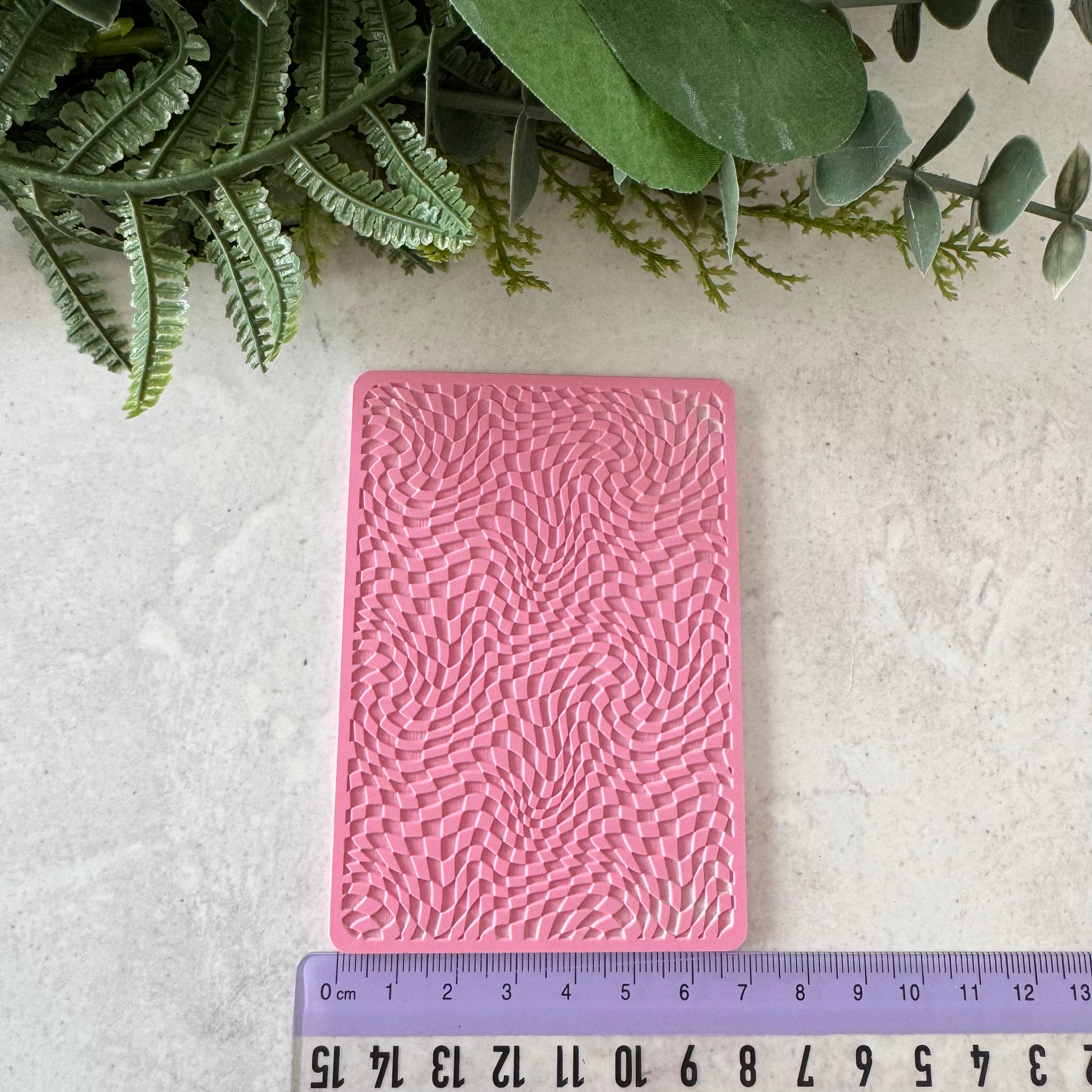 Pink embossed rectangular object on a light gray surface with green plants in the background