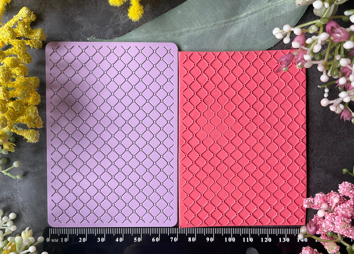 Two rectangular cards with a raised geometric lattice pattern, one lavender and one coral pink, displayed side-by-side on a dark slate background with a black ruler at the bottom and floral decorations in the corners.